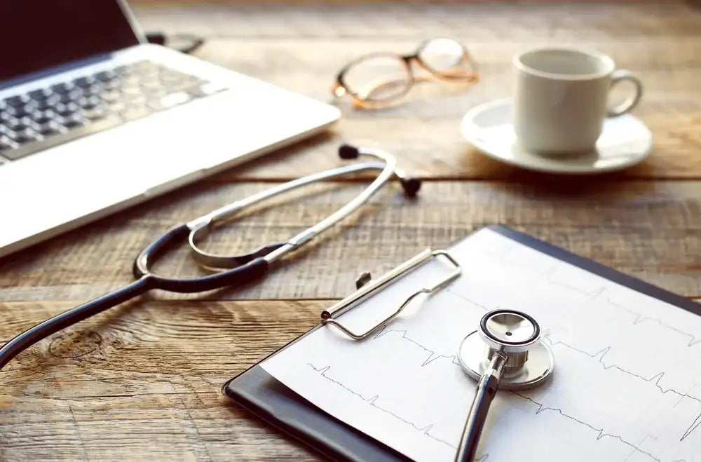 Stethoskop auf einem Klemmbrett mit EKG-Kurven, daneben Laptop, Brille und Kaffeetasse auf einem Holztisch.