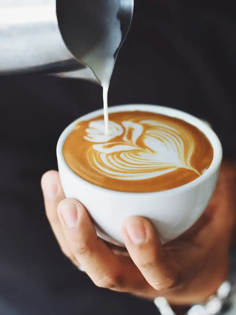 Milchschaum wird aus einem Kännchen in eine weisse Tasse gegossen und formt ein Latte-Art-Muster auf dem Kaffee.